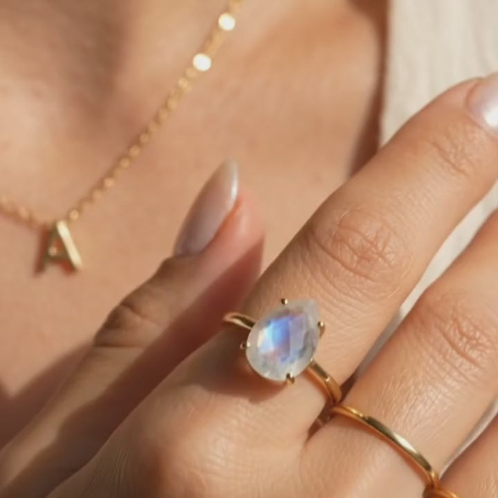 A video showcasing a Moonstone teardrop ring in yellow gold with pear rainbow moonstone on warm neutral fabric