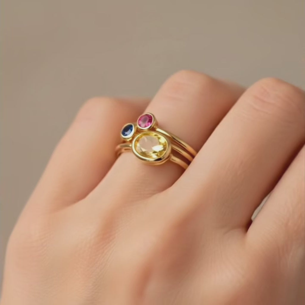 A tiny ruby ring is shown with an open back and rounded band. A panover shows a set of silver, rose gold, and gold rings in open boxes A hand in the corner wears a gold stack of mixed birthstone rings. 