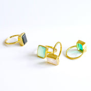 Four Large Rectangle Gemstone Rings in Vermeil Gold Lined up diagonally on a white background. Black Onyx Rectangle Ring, Green Emerald Quartz May Birthstone Ring, Aqua Chalcedony March Birthstone Ring, and Rainbow Moonstone June Birthstone Ring