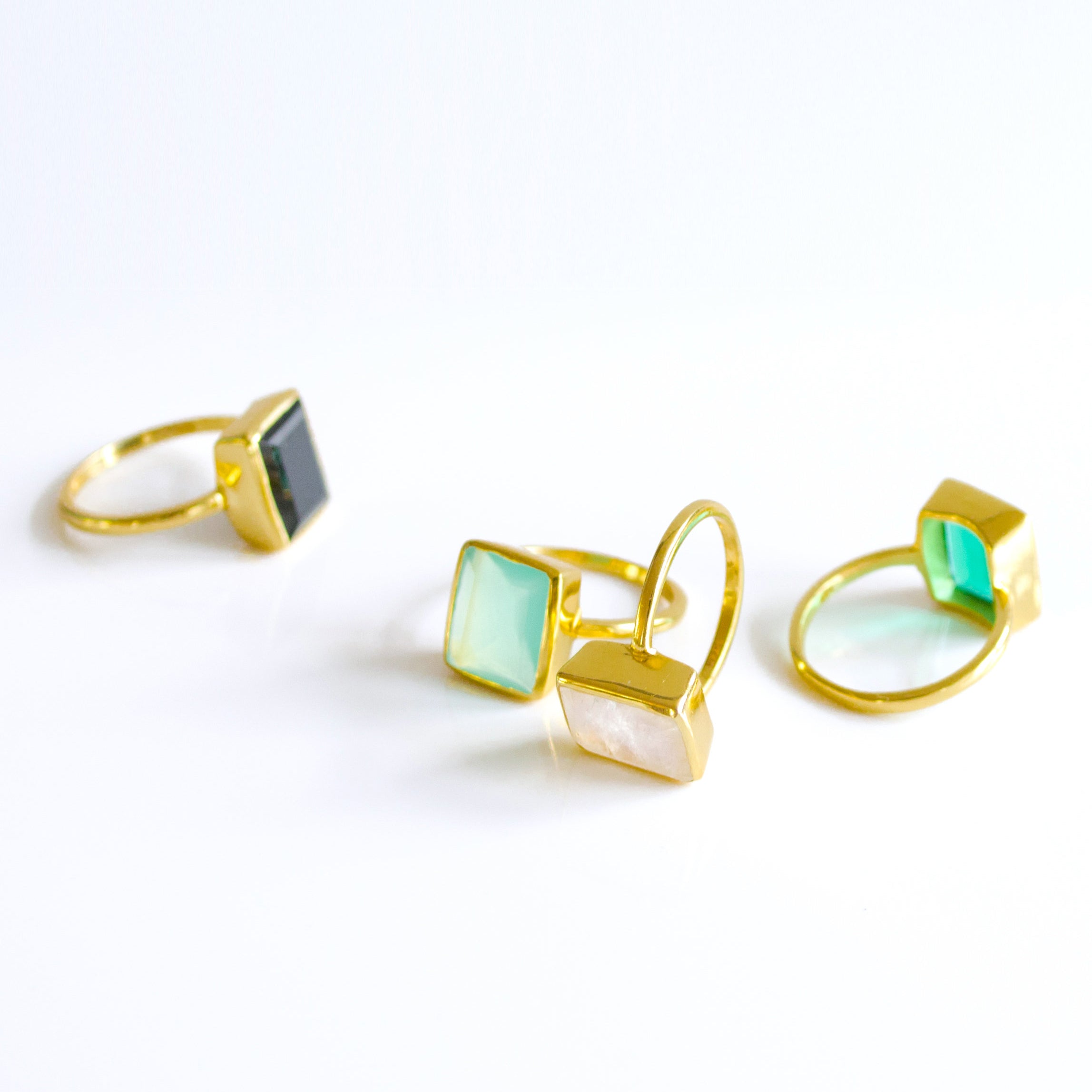 Four Large Rectangle Gemstone Rings in Vermeil Gold Lined up diagonally on a white background. Black Onyx Rectangle Ring, Green Emerald Quartz May Birthstone Ring, Aqua Chalcedony March Birthstone Ring, and Rainbow Moonstone June Birthstone Ring
