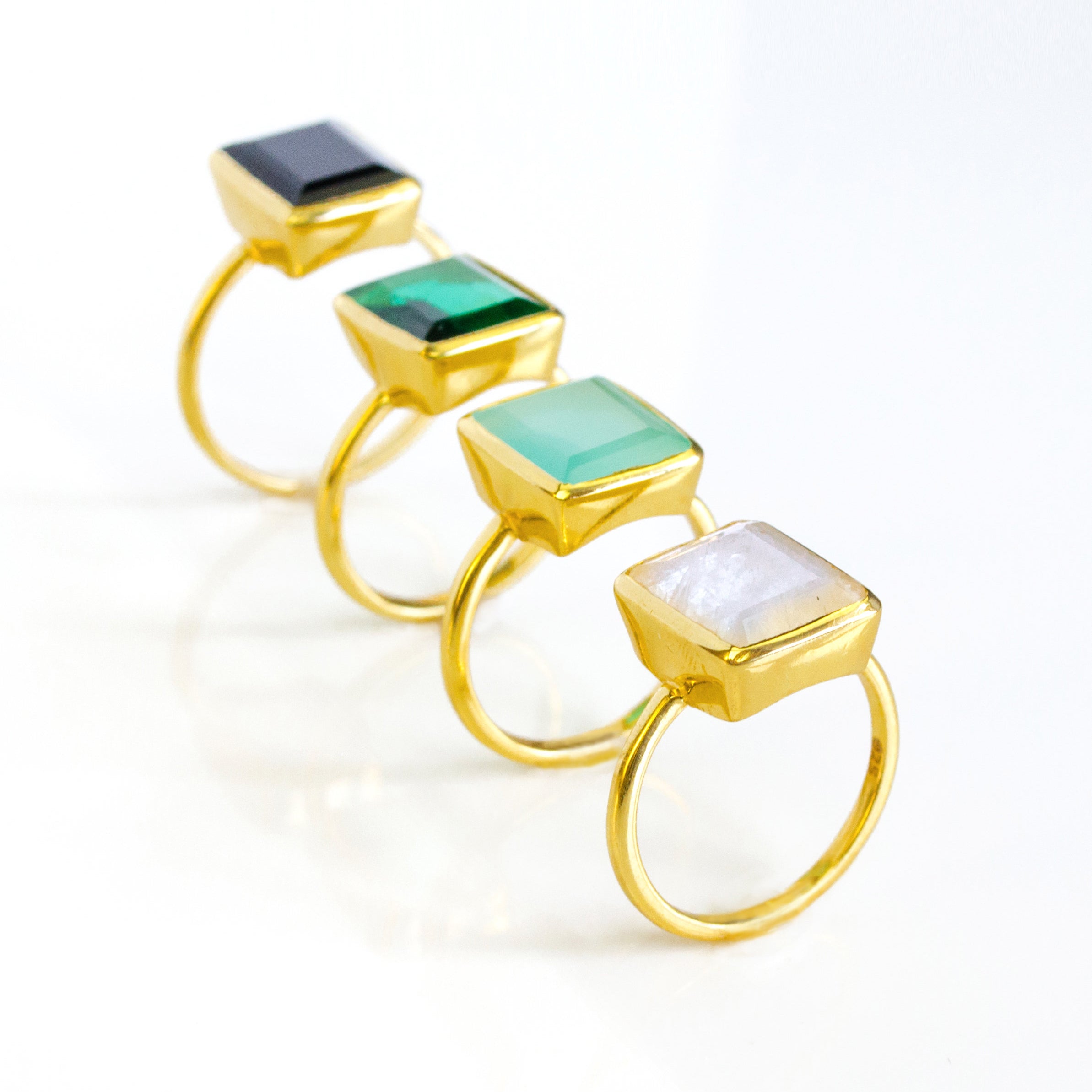 Four Large Rectangle Gemstone Rings in Vermeil Gold Lined up diagonally on a white background. Black Onyx Rectangle Ring, Green Emerald Quartz May Birthstone Ring, Aqua Chalcedony March Birthstone Ring, and Rainbow Moonstone June Birthstone Ring