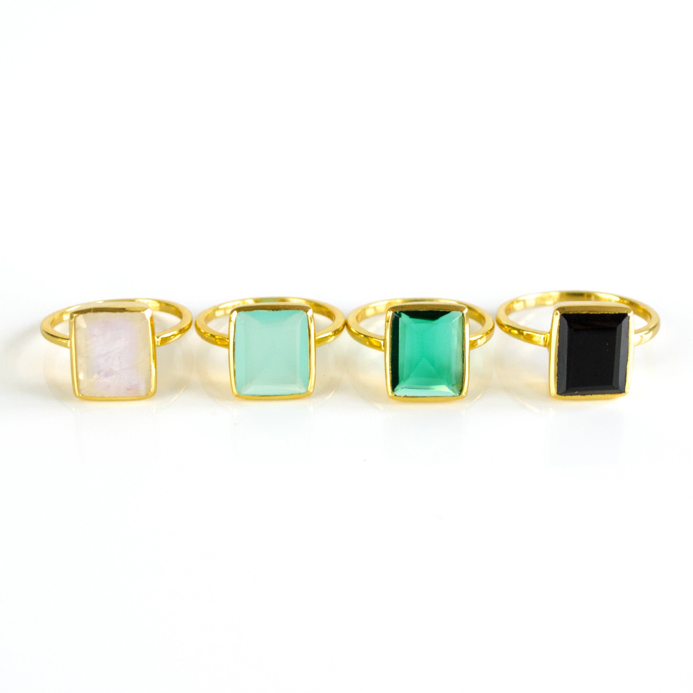 Four Large Rectangle Gemstone Rings in Vermeil Gold Lined up diagonally on a white background. Black Onyx Rectangle Ring, Green Emerald Quartz May Birthstone Ring, Aqua Chalcedony March Birthstone Ring, and Rainbow Moonstone June Birthstone Ring