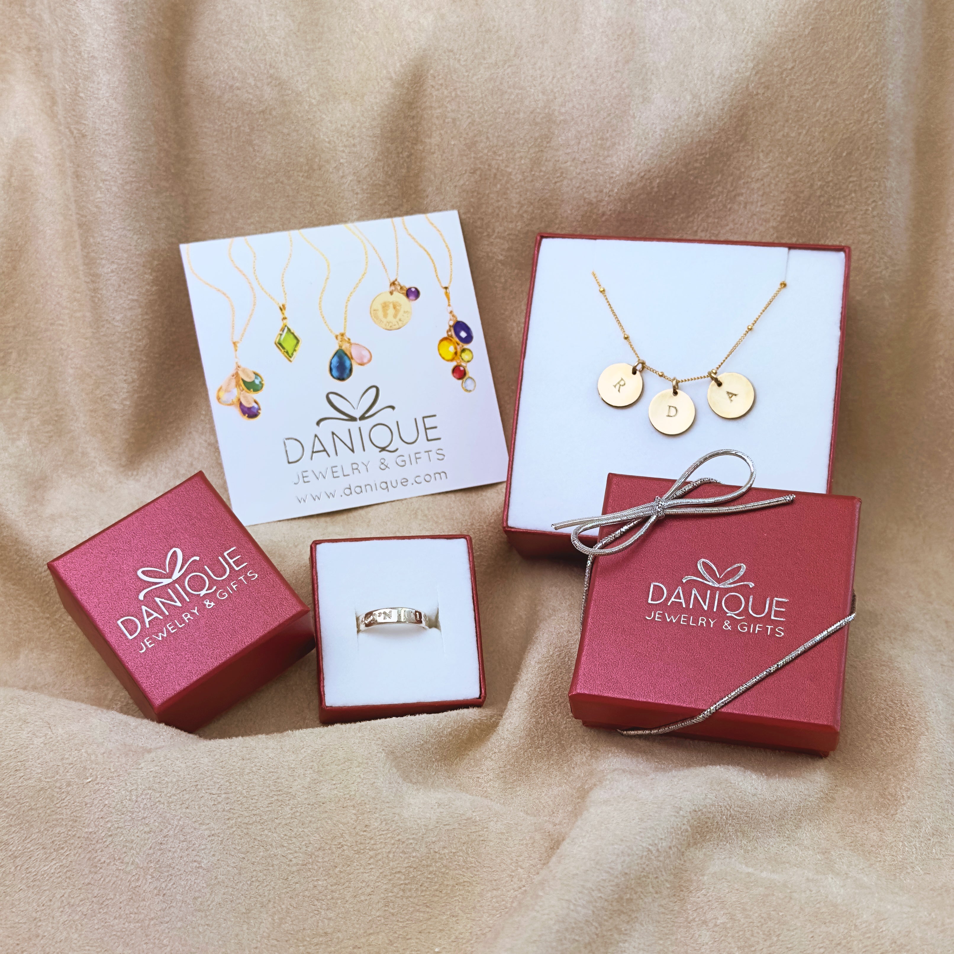 2 red Danique Jewelry boxes on a velvet background shows a gold initial disc necklace in one box and a silver engraved ring in another box. A Danique Jewelry post card sits in the upper left corner of the box setup.