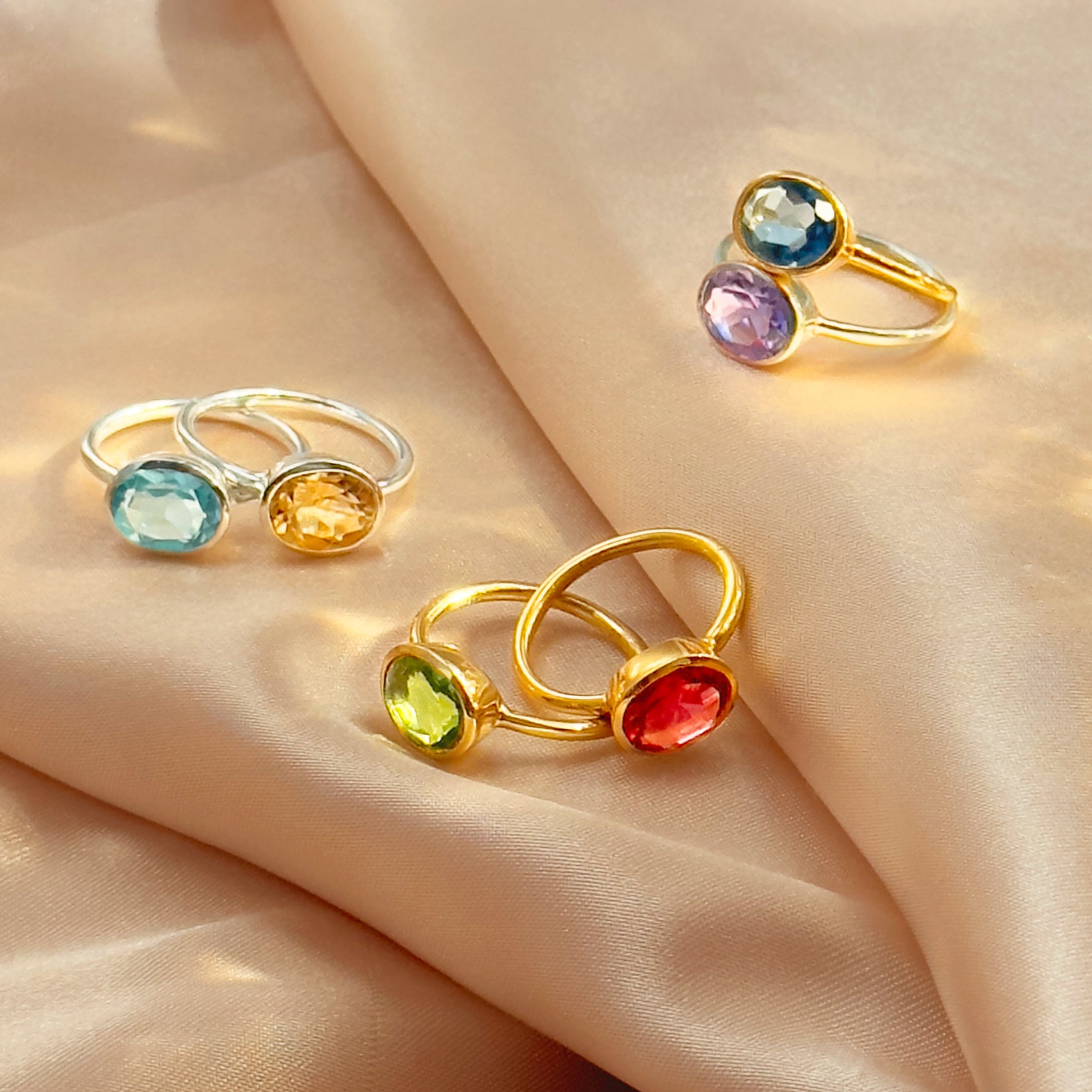 Assorted oval birthstone rings arranged on blush satin—bezel-set ovals on yellow-gold tone and polished sterling-silver bands. Build a colorful ring stack or bold statement ring look with gemstones such as aquamarine (March), citrine (November), peridot (August), ruby red (July), and amethyst (February). A modern collection of cocktail rings for everyday style or anniversary jewelry for her.