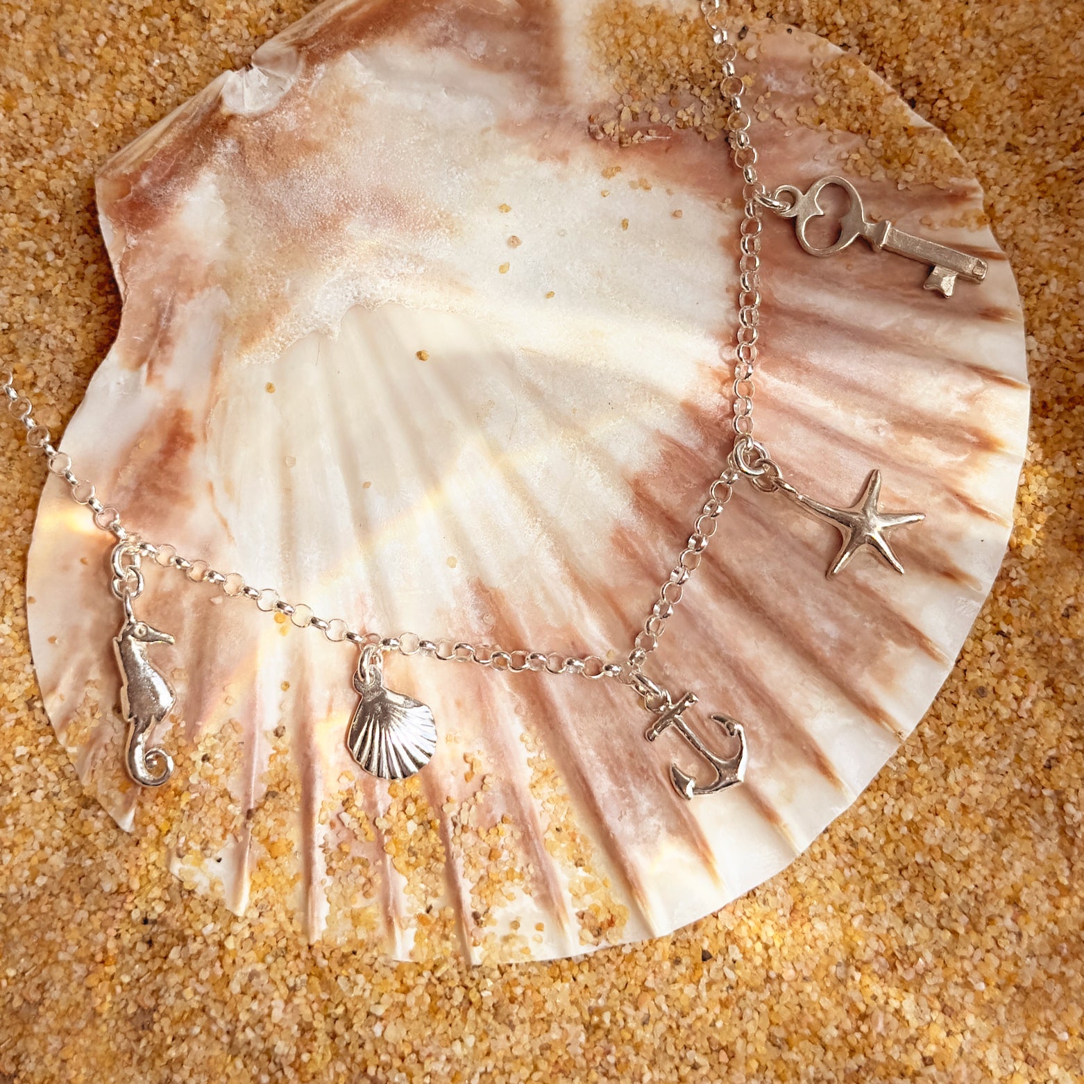 Sterling silver ocean-themed charm anklet with seahorse, seashell, anchor, starfish, key, and heart charms displayed on a scallop shell with beach sand background – perfect personalized summer gift idea. custom charm anklet, build your own anklet, sterling silver anklet, summer anklet jewelry, personalized beach anklet, ocean charm anklet, charm anklet gift, handmade anklet, beach vacation jewelry, coastal jewelry for her, best friend summer gift, custom anklet for her, friendship anklet gift, beach wedding