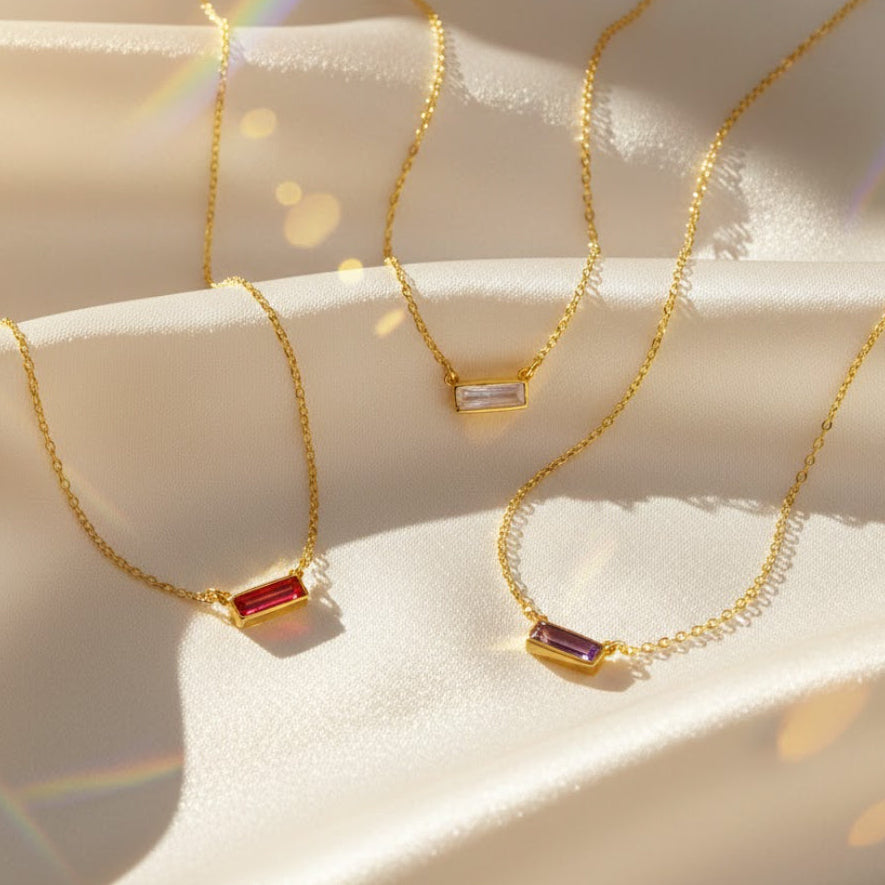 Tiny Adira birthstone bar necklace in 18k gold vermeil displayed on soft satin, featuring petite faceted gemstone bars in multiple birthstones including ruby, citrine, amethyst, aquamarine and more, shown with delicate cable chains. A minimalist personalized jewelry piece available in all twelve birthstones with optional hidden engraving, perfect for bridesmaids, birthdays, anniversaries, and everyday gifting for women who love dainty gemstone necklaces.