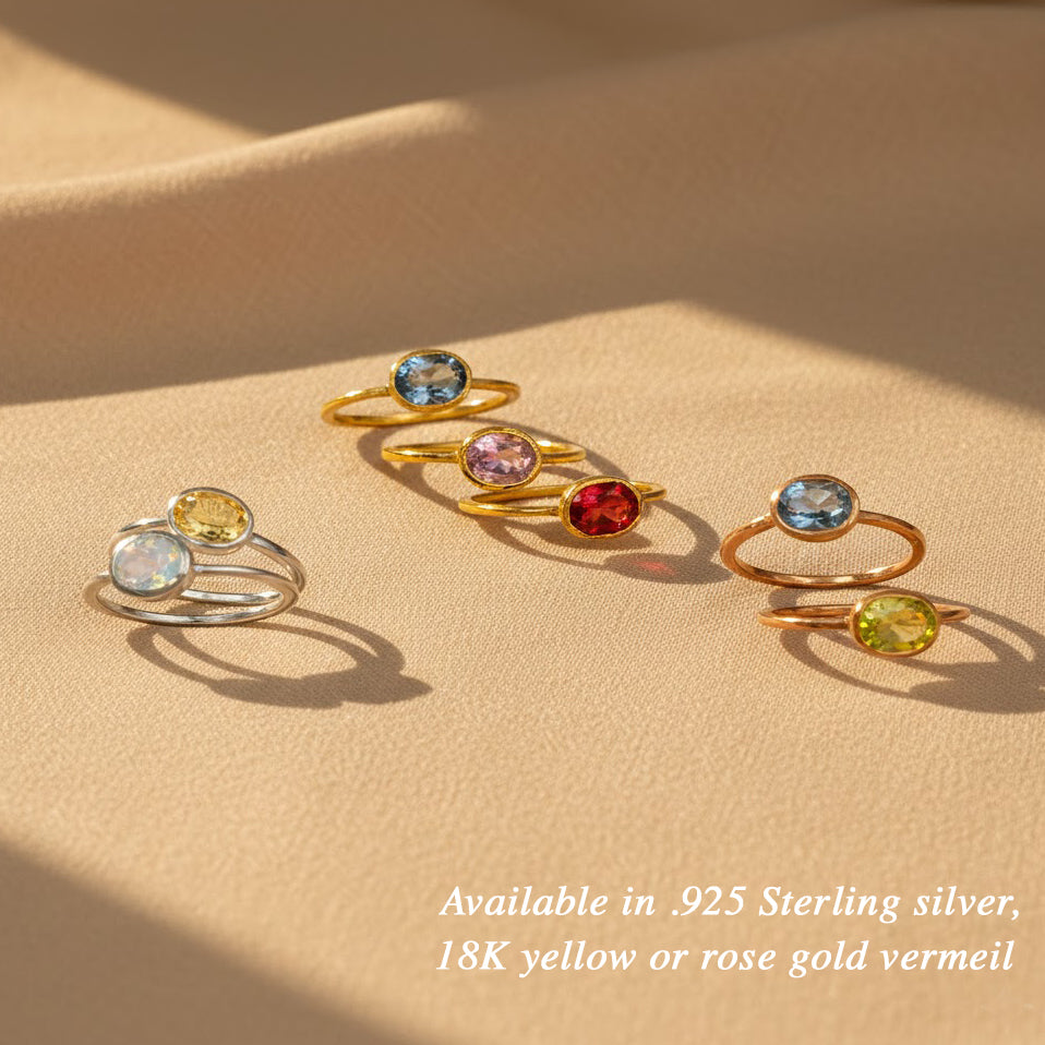 Flat lay of stacking birthstone rings arranged on neutral fabric, colorful oval January Garnet, March Aquamarine, April Diamond Clear Quartz in gold and silver bands, minimalist birthstone ring collection for women who love layered jewelry