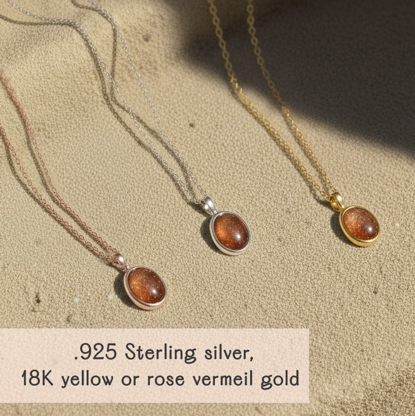 Dainty oval sunstone necklaces in .925 sterling silver, 18k yellow gold vermeil and 18k rose gold vermeil displayed on a warm sand textured background, each pendant featuring a glowing coppery orange sunstone cabochon with subtle sparkle on a fine minimalist chain. Elegant boho gemstone necklace perfect for everyday wear or layering, a meaningful gift for her, mom, sister or best friend for birthdays, anniversaries, Christmas or Mother’s Day.