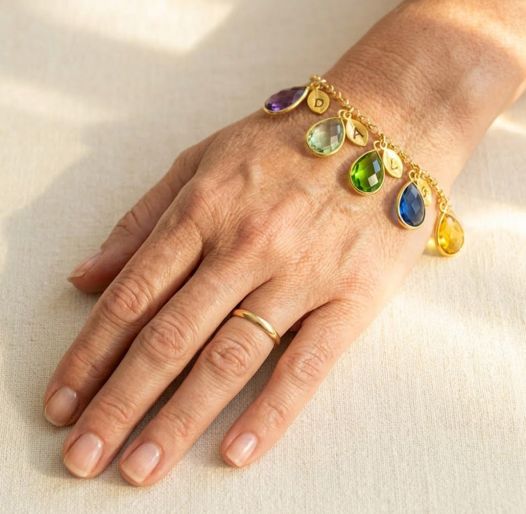 Custom birthstone and initial charm bracelet shown on hand, personalized family bracelet in gold