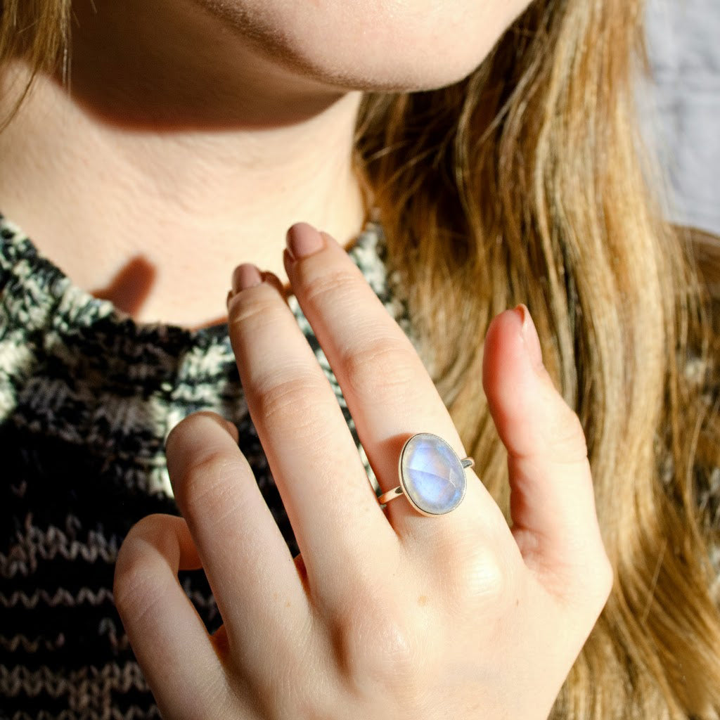 Lifestyle image of a woman wearing a rainbow moonstone oval ring, showcasing the soft glow of a bezel-set June birthstone. Minimalist gold moonstone ring styled for everyday elegance, stacking, and effortless fashion. Thoughtful gemstone jewelry gift for her, featuring a timeless oval silhouette and handcrafted design.