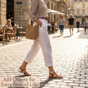 Person walking on a cobblestone street with a woven handbag, wearing white pants and a sterling silver personalized charm anklet with sandals.