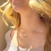 A person with blond hair wears a 5 stone horizontal sterling silver birthstone bar necklace. Custom bezel set birthstone pendant for mom with kids birthstone. Minimalist family tree jewelry for mom.