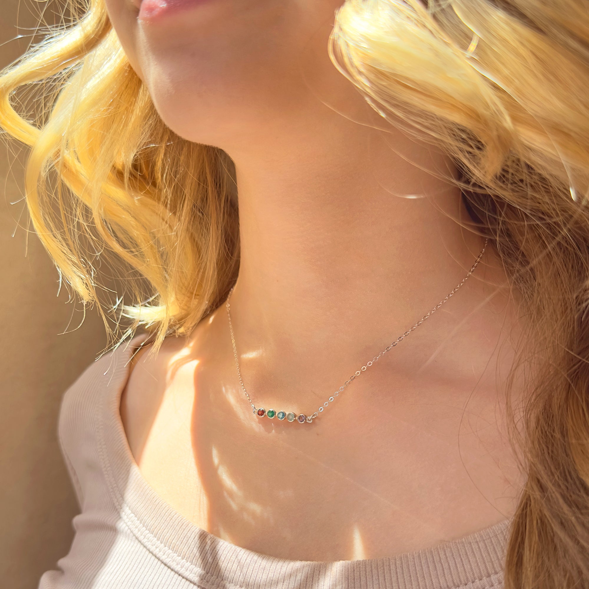 A person with blond hair wears a 5 stone horizontal sterling silver birthstone bar necklace. Custom bezel set birthstone pendant for mom with kids birthstone. Minimalist family tree jewelry for mom.
