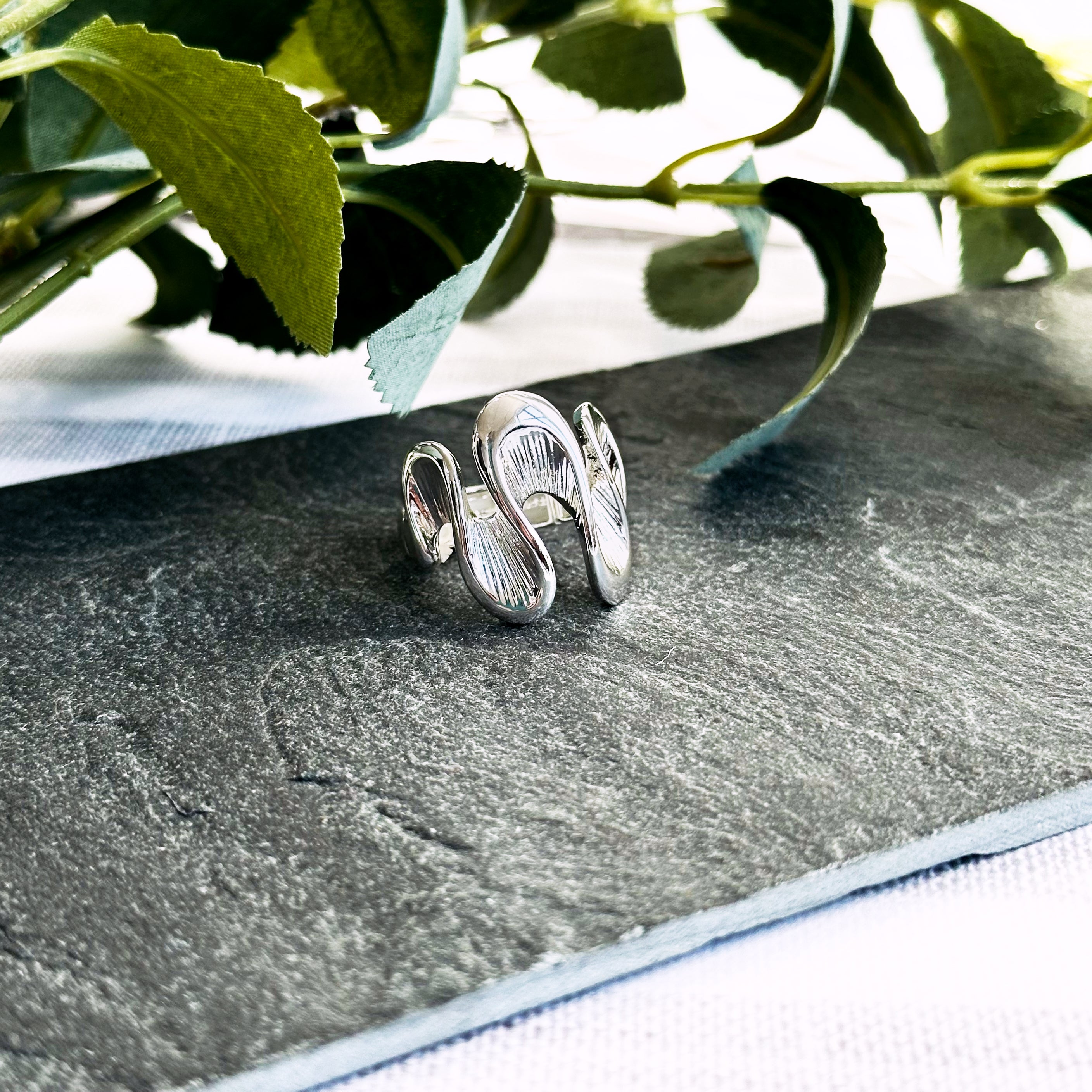 Silver Waves ring rests on a flat dark stone with leaves in the background, Model wears silver textured wave shaped ring on index finger. Stretchy ring, adjustable to fit ring, wave ring, nature inspired jewelry, boho jewelry, nature inspired ring, ocean ring, water ring, bohemian jewelry, zinc alloy, gold, silver, one size fits most, ring for her, mother's day git, christmas gift, gift for girlfriend