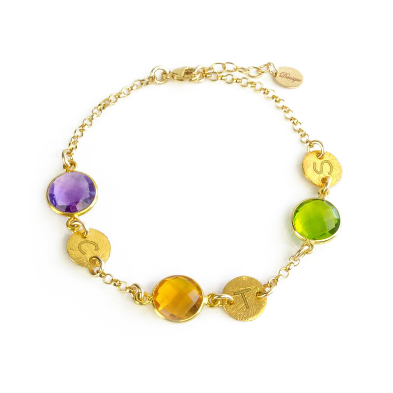 A gold bracelet featuring peridot and citrine and purple amethyst birthstones rests on a white background. Each birthstone is accompanied by a round disc charm with an initial hand stamped into it. August birthstone and September birthstone, green gemstone, purple gemstone, yellow gemstone, custom made personalized mother's bracelet with birthstones