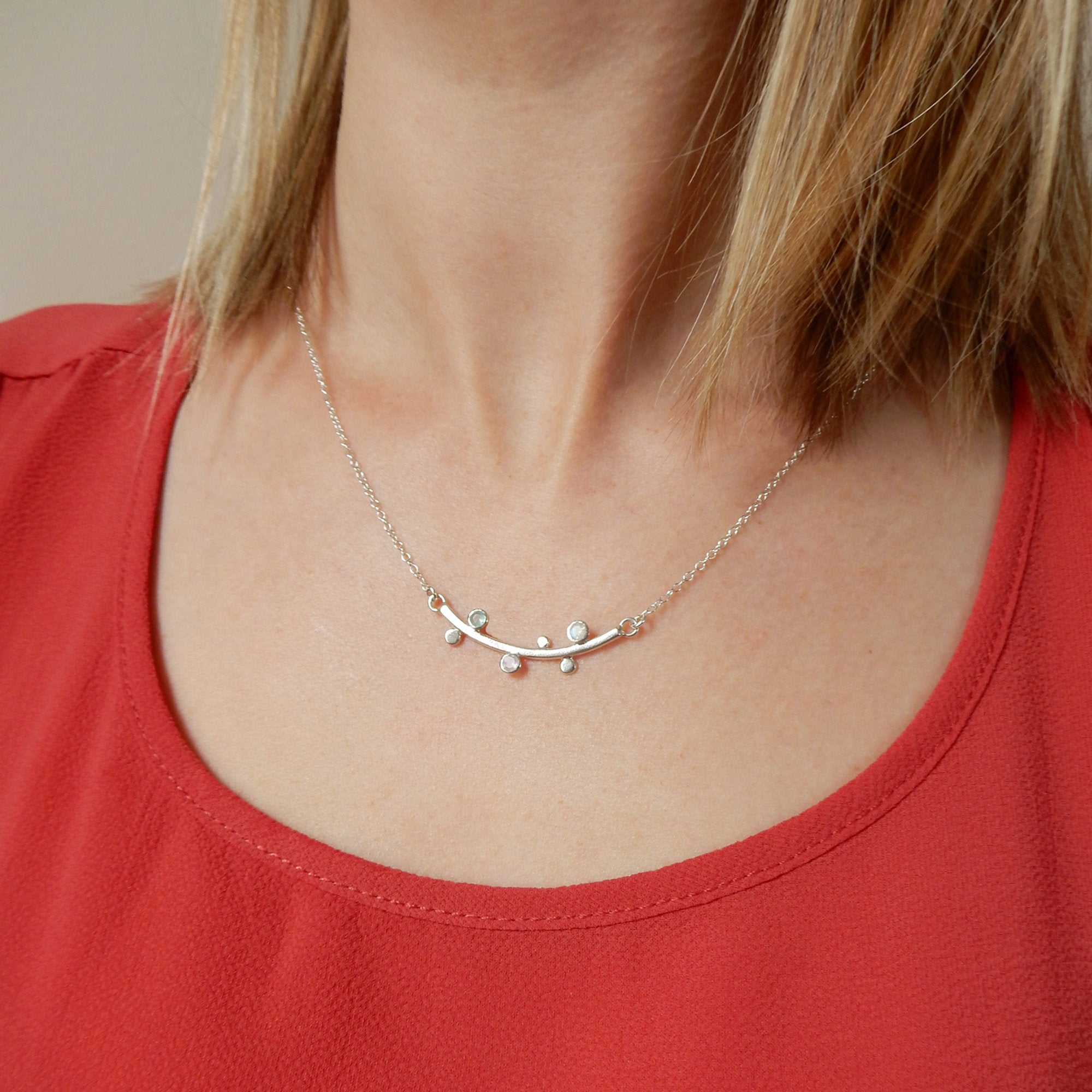 A person with shoulder length blonde hair wears a silver curved bar necklace with inlaid bezel set small dainty birthstones. This is the perfect Mother's Day, Christmas or Birthday gift for mom or grandma with her children's or grandchildren's birthstones carefully hand set into each piece. Available in 925 sterling silver, 18k gold vermeil or 18k rose gold vermeil. Necklace can be made to any length needed. 