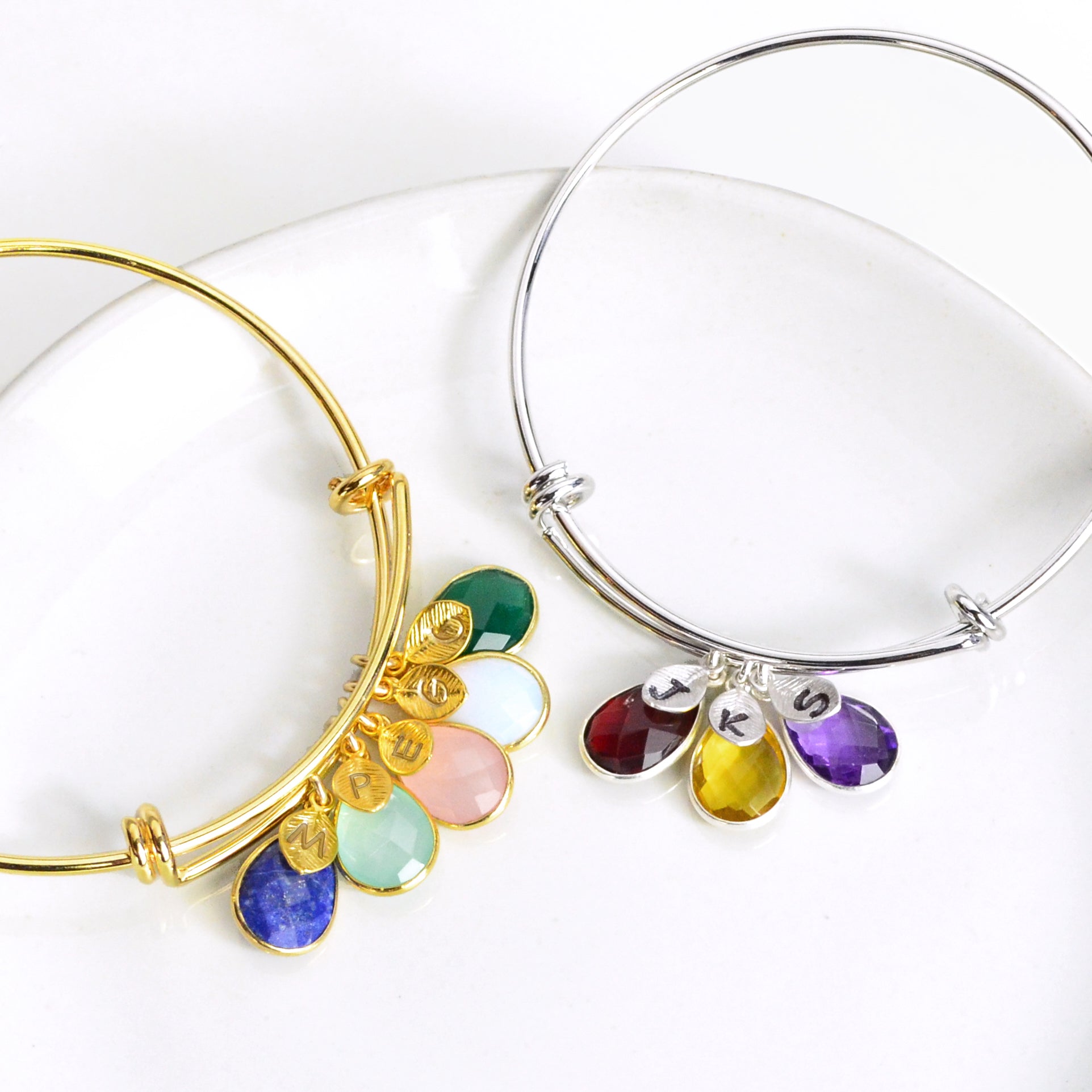 Two bangle birthstone bracelets on display. Single gold bangle bracelet with stamped leaf charms and lapis, aqua chalcedony, piny chalcedony, opalite and green onyx birthstones. Single silver bangle with hand stamped leaf charms and ruby, citrine and purple amethyst birthstones. 