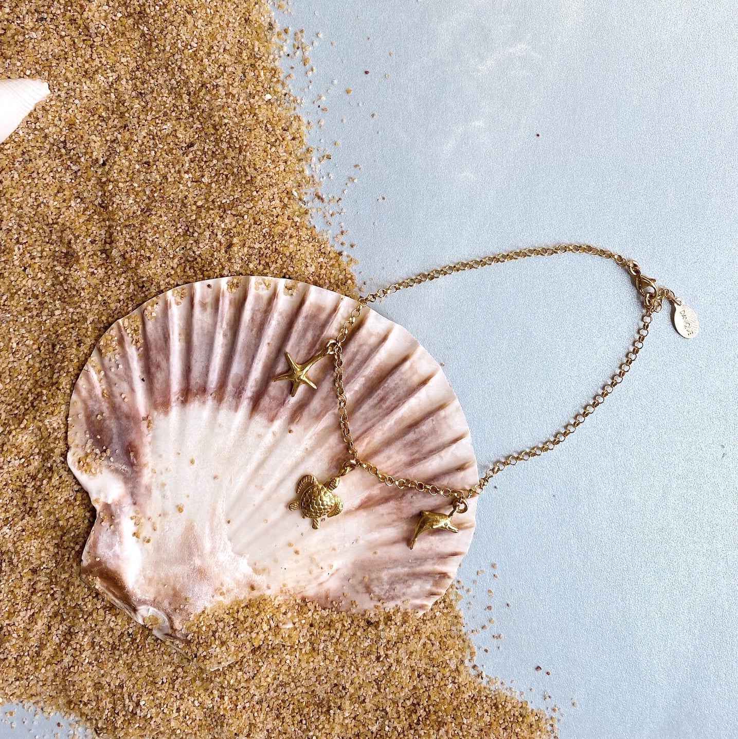 Single gold filled charm anklet with seashell, turtle and dolphin charms and lobster clasp sitting in a seashell and sand. 