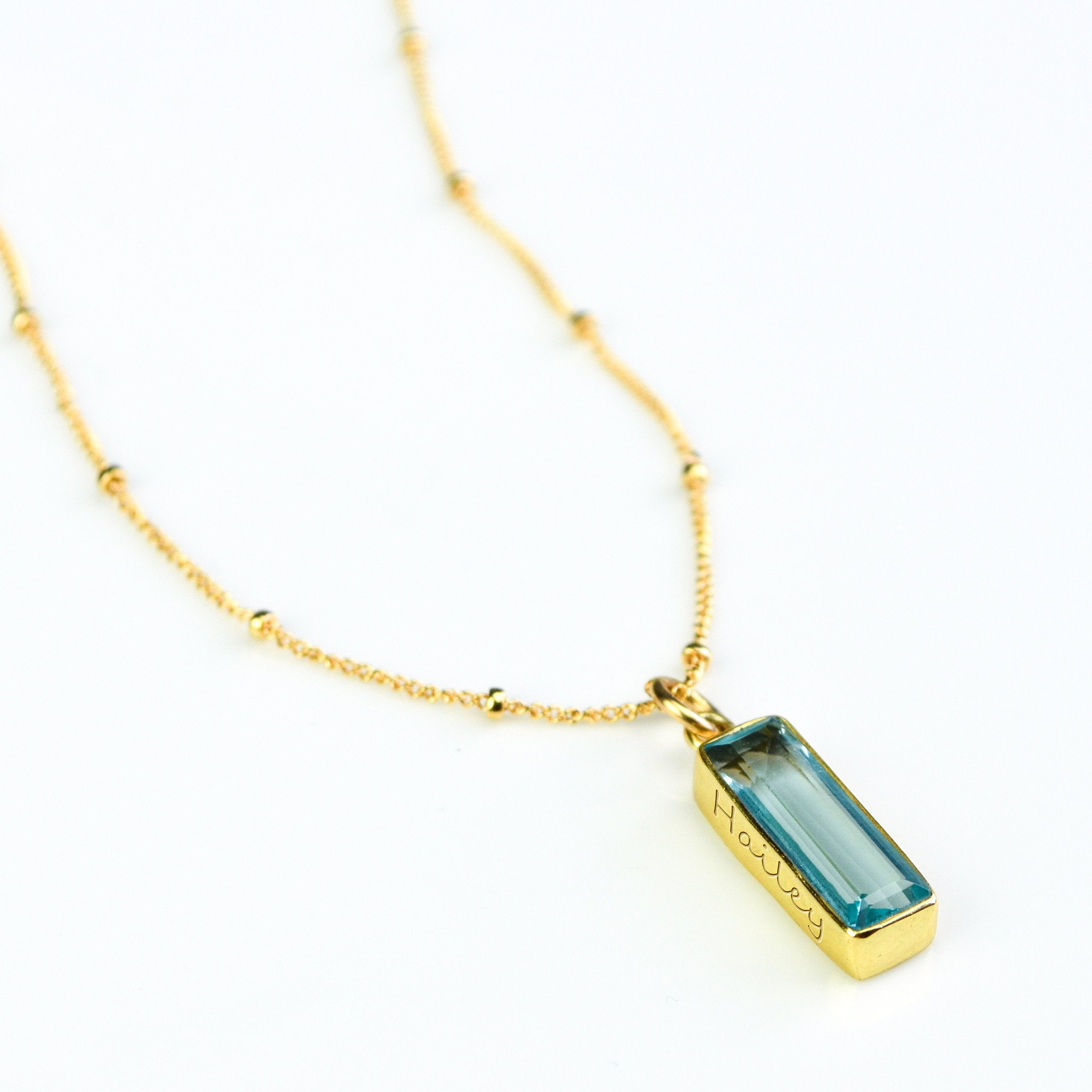 Single gold filled satellite chain with vertical sky blue gemstone with secret message engraved december birthstone adira bar necklace on white background