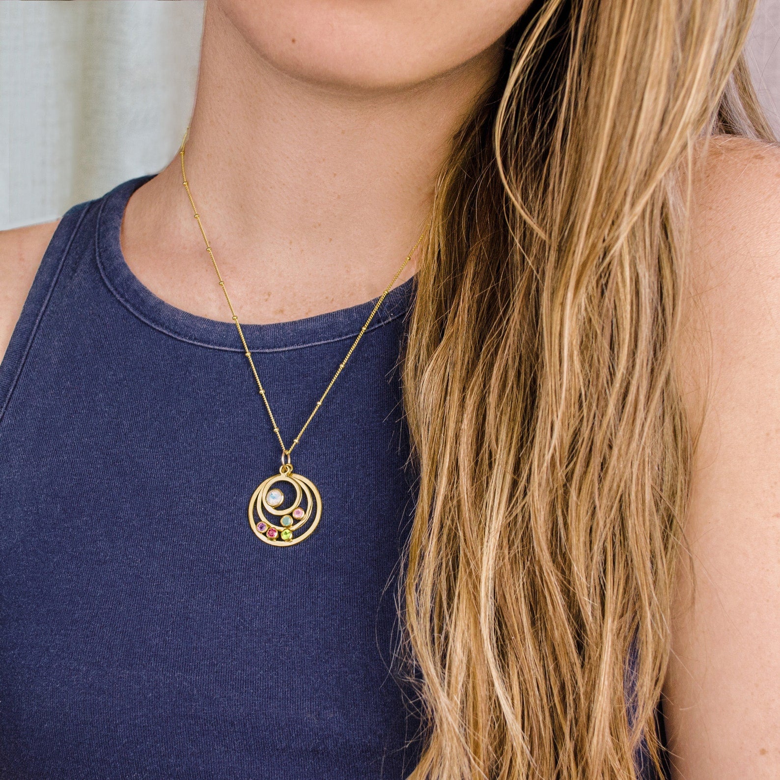 Model in blue top is wearing a gold filled satellite chain with three orbits featuring moonstone, aqua chalcedony, pink chalcedony, purple amethyst, ruby and peridot birthstones. 