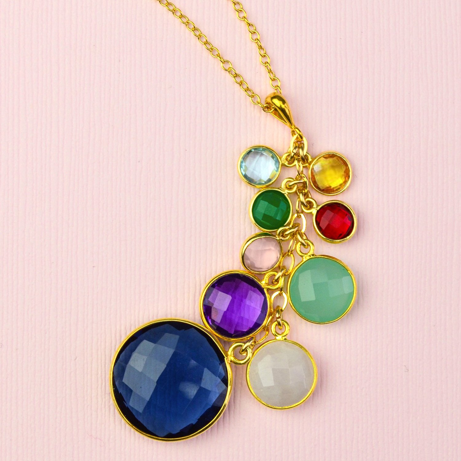 A gold grandmother cascade necklace lays against a pink background. September birthstone necklace for grandma, blue topaz, citrine, green onyx, ruby, pink chalcedony, aqua chalcedony, amethyst, moonstone. Vermeil Gold Gold-Filled Chain Grandmother Necklace with children & grandchildrens birthstones round gemstones