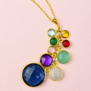 A gold grandmother cascade necklace lays against a pink background. September birthstone necklace for grandma, blue topaz, citrine, green onyx, ruby, pink chalcedony, aqua chalcedony, amethyst, moonstone. Vermeil Gold Gold-Filled Chain Grandmother Necklace with children & grandchildrens birthstones round gemstones
