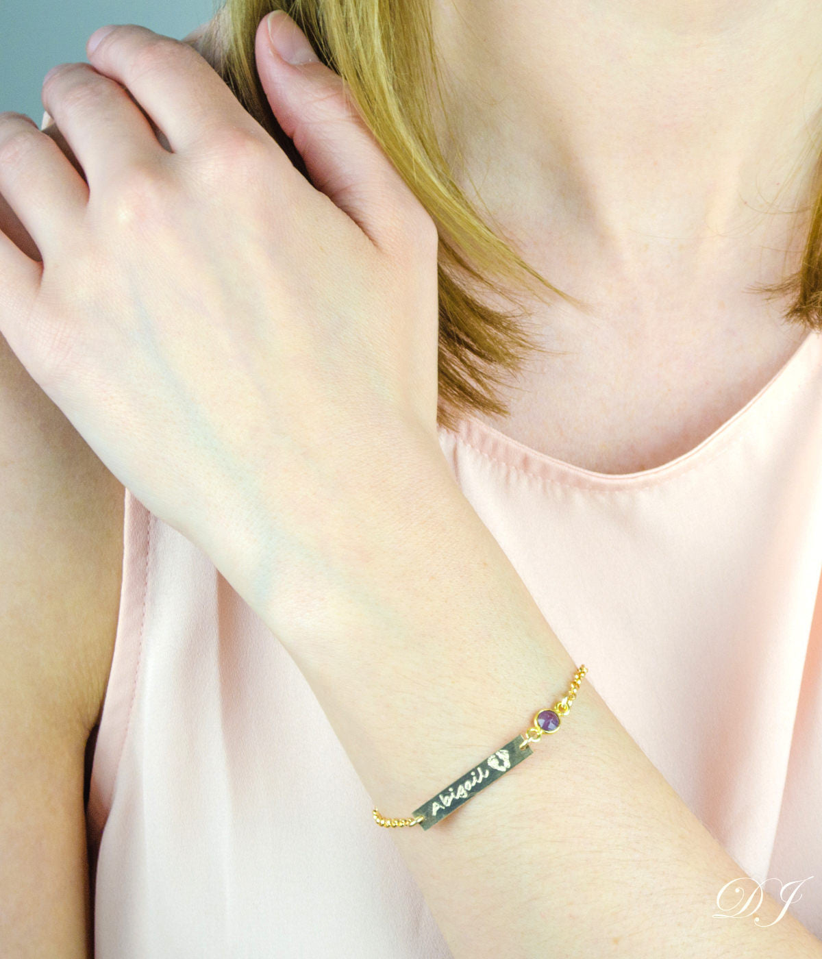Model in pink top is wearing a gold filled footprint name engraved bar bracelet with purple amethyst gemstone. 