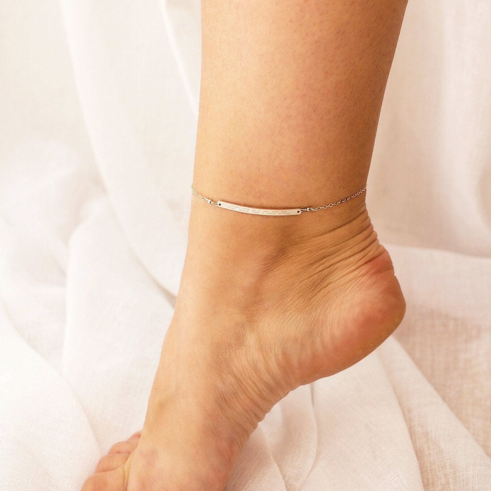Model is sporting a single silver cable chain morse code bar anklet. 