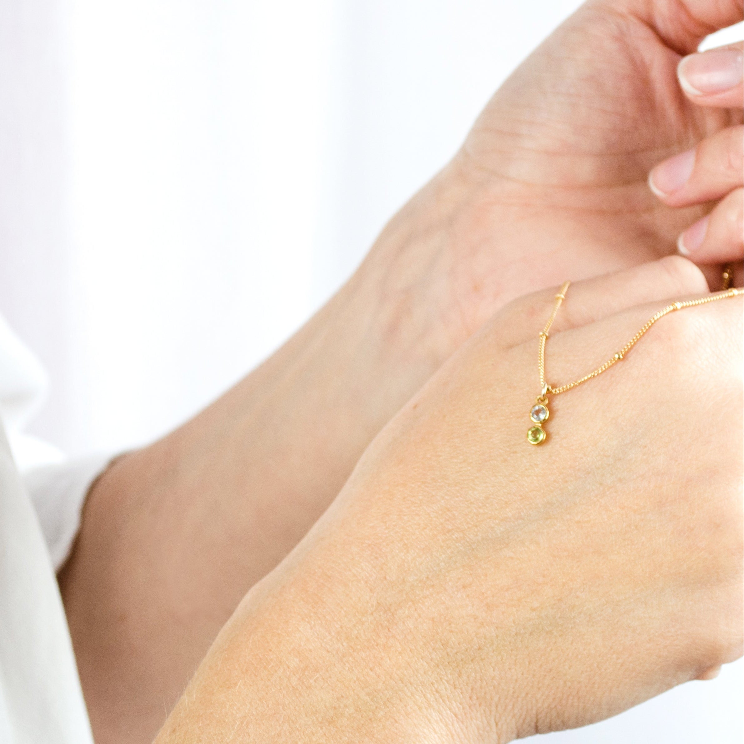 Model is holding a gold filled satellite chain with 2 birthstones on the vertical bar necklace including peridot and clear quartz, the birthstones for August and April respectively.