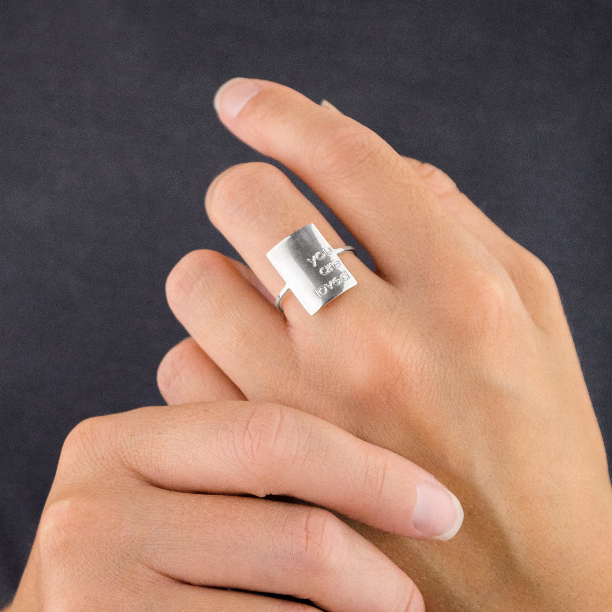 Model is wearing a sterling silver rectangular name plate ring with the phrase you are loved engraved. 
