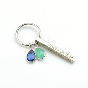 Minimal product view of a silver mom keychain photographed on a bright white background. A dainty 3D bar is engraved with a name, paired with two bezel-set birthstones in ocean hues. Lightweight and durable for daily use, this personalized keychain can include additional stones for siblings and custom text on up to four sides for a heartfelt, practical gift.