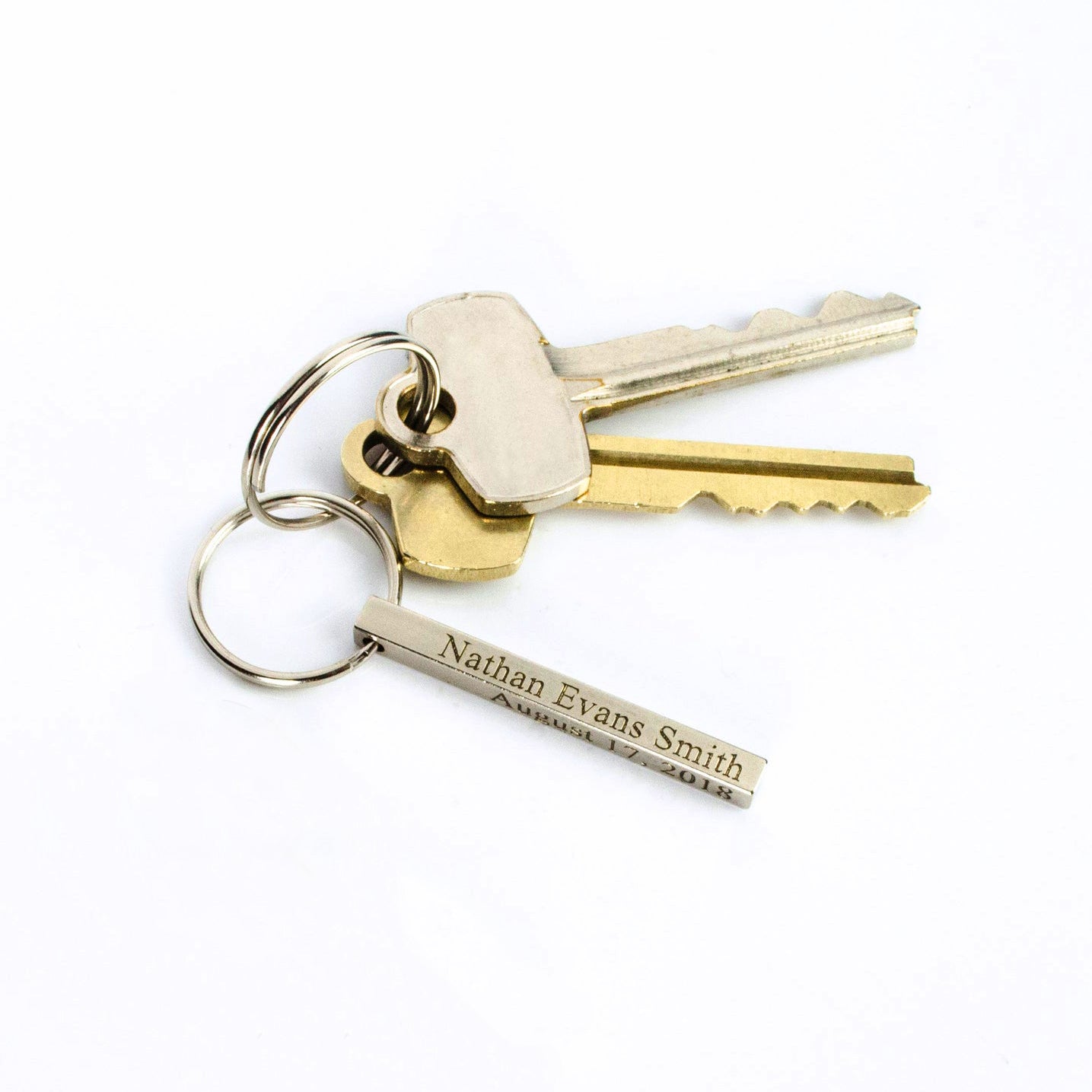 Minimal silver small 3D bar keychain attached to a set of keys on a bright white background. The compact 40×4 mm bar shows fine laser lettering and smooth corners, designed for pockets without bulk. Customize this personalized keychain with monogram initials, roman numerals, or latitude and longitude to create a meaningful everyday accessory for him or her.